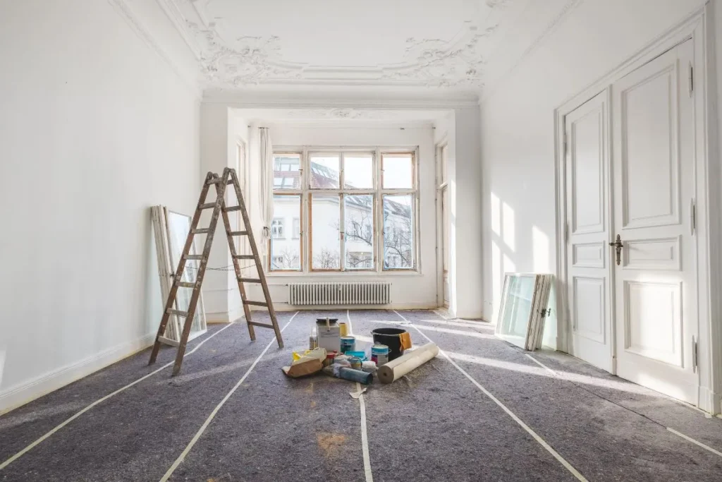 Bright, empty room under renovation with a vintage ladder, paint cans, and materials on a carpeted floor. Sunlight streams through large windows.
