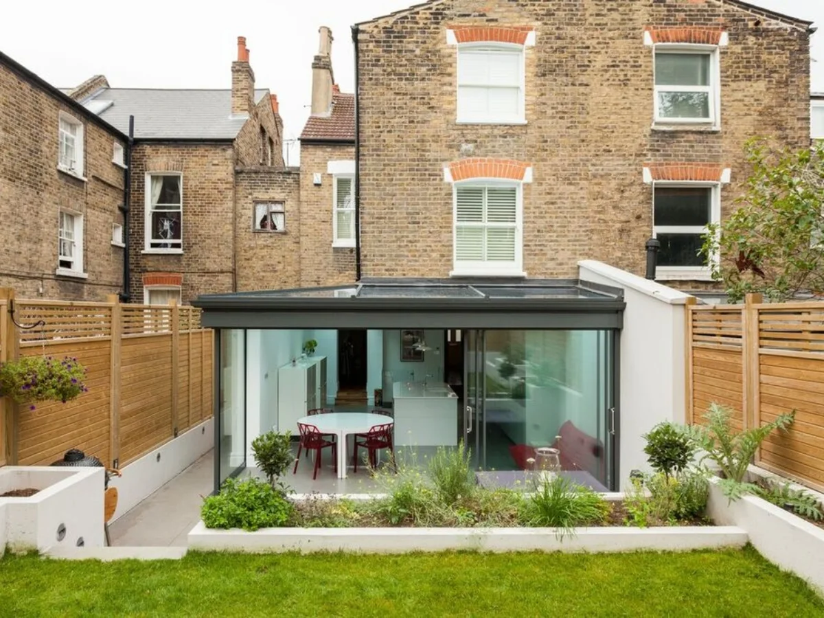 wrap- around extension A modern glass extension sits behind a historic brick townhouse, with dining furniture visible inside and a tidy green garden outside; calm and inviting.